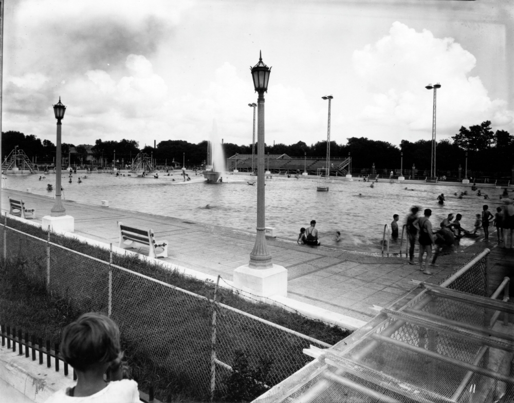 Black and white photo of a crowded public swimming pool surrounded by tall lights and benches. A fountain sprays water in the pools center. People are swimming, lounging, and walking along the poolside. A fence and a cloudy sky are visible.