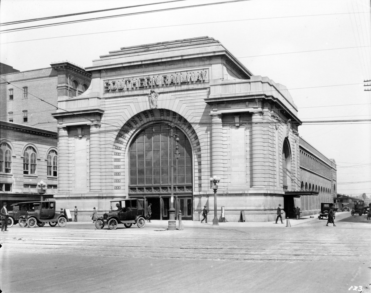 A historic black-and-white photo of a grand railway station entrance with Southern Railway on the arch. Vintage cars and pedestrians populate the scene, showcasing early 20th-century urban life.