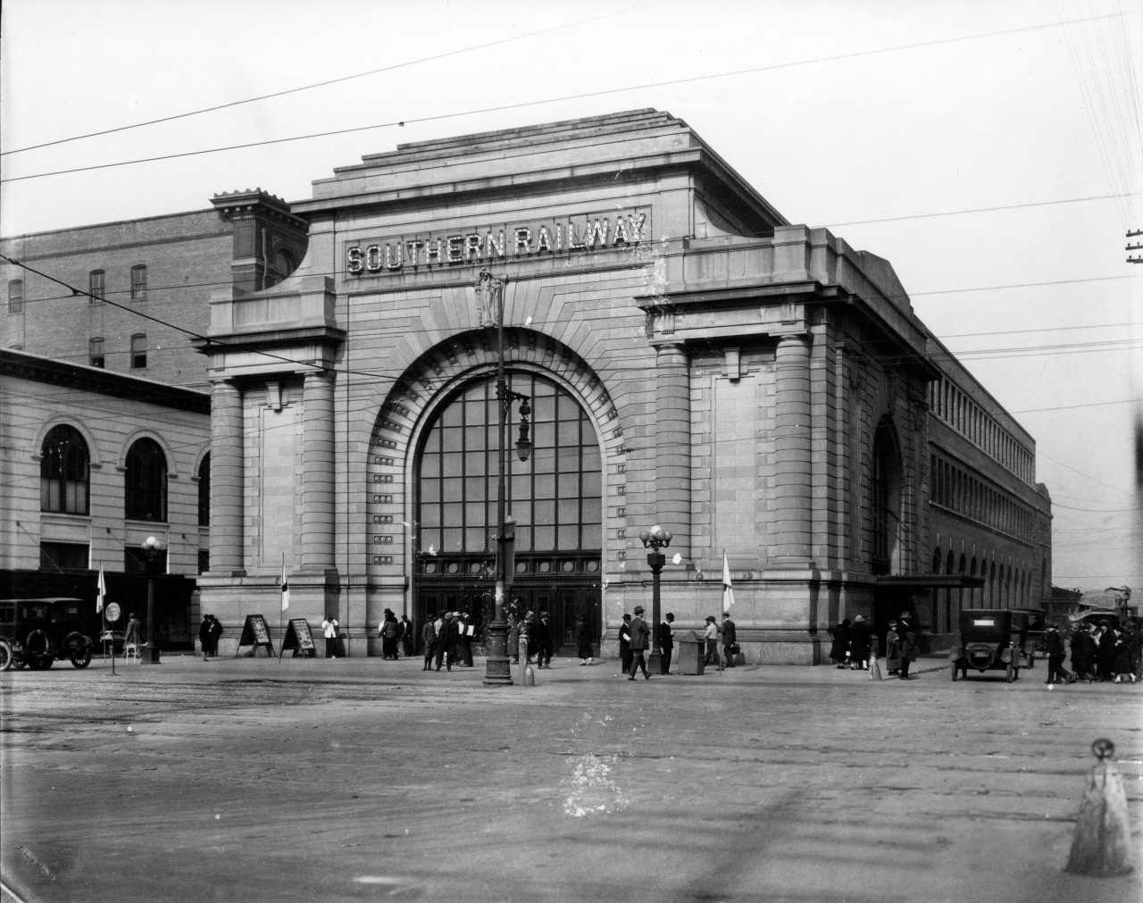 A historic black-and-white photo of a large train station with Southern Railway written above a grand arched entrance. People and vintage cars are gathered in the surrounding area.