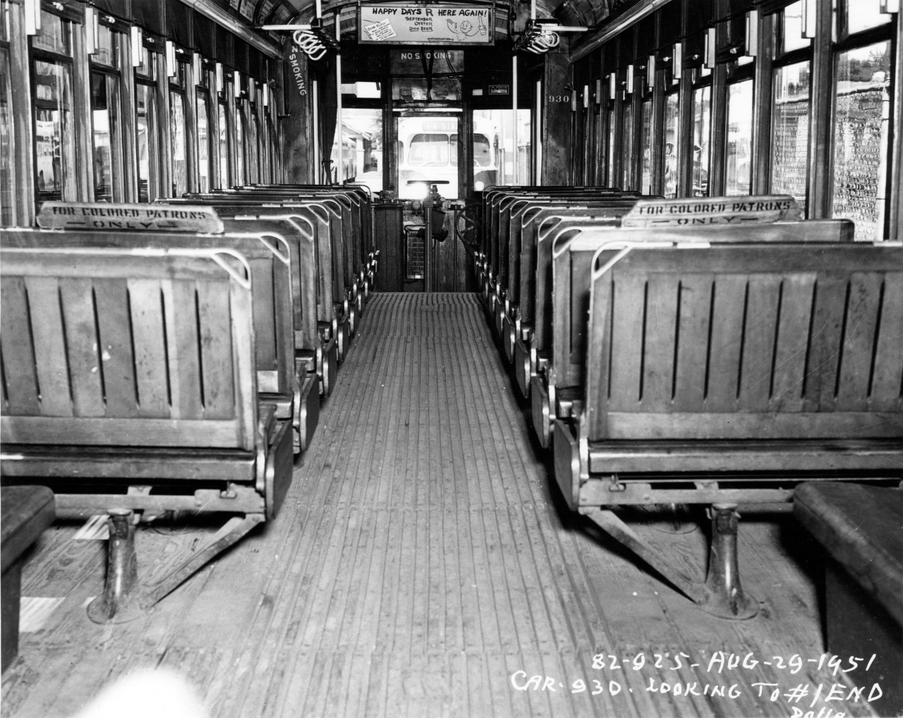 Black and white photo of a vintage streetcar interior with wooden benches in rows on both sides. Signs above seats read For Colored Patrons. The front of the car displays the date Aug. 29, 1951 and car number 930.