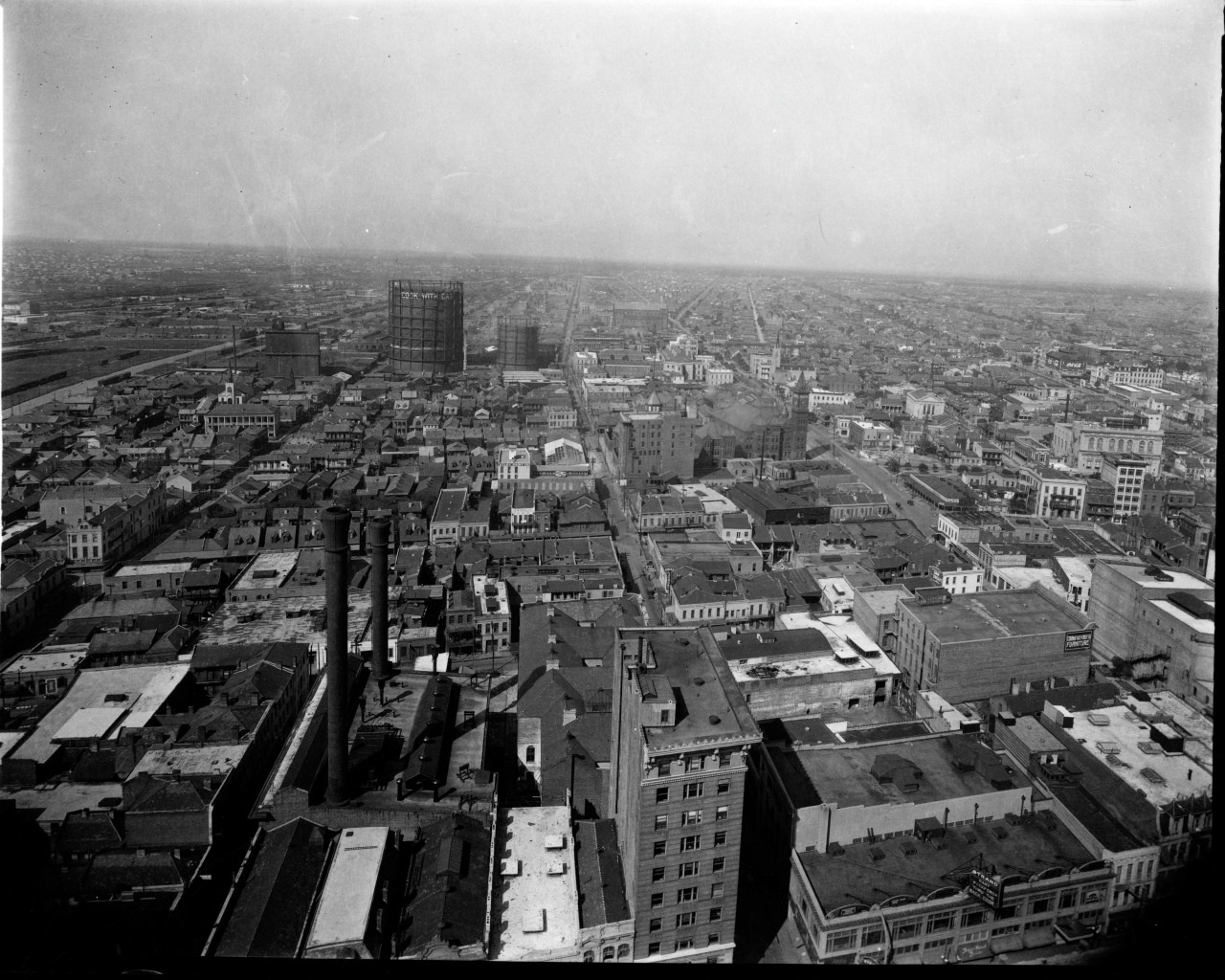 A historic black-and-white aerial view of a sprawling urban landscape, featuring densely packed buildings, factories with smokestacks, and wide streets. The horizon is visible in the distance under a clear sky.