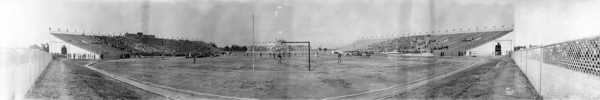 Panoramic view of an early 20th-century sports stadium, featuring a large field surrounded by two grandstands filled with spectators. Several athletes are visible on the field, and flags line the top of the bleachers.