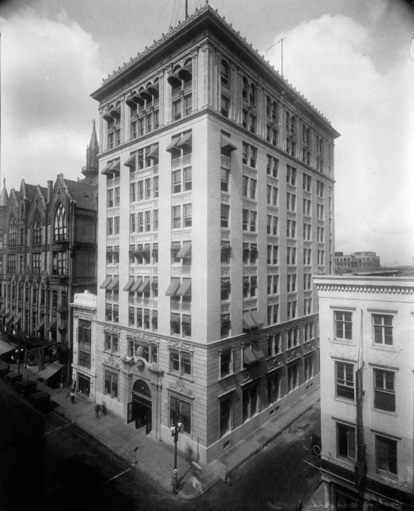 A historic black-and-white photograph of a large multi-story building with intricate architectural details, including arched windows and decorative elements. It is situated on a street corner next to a smaller building and opposite a church.