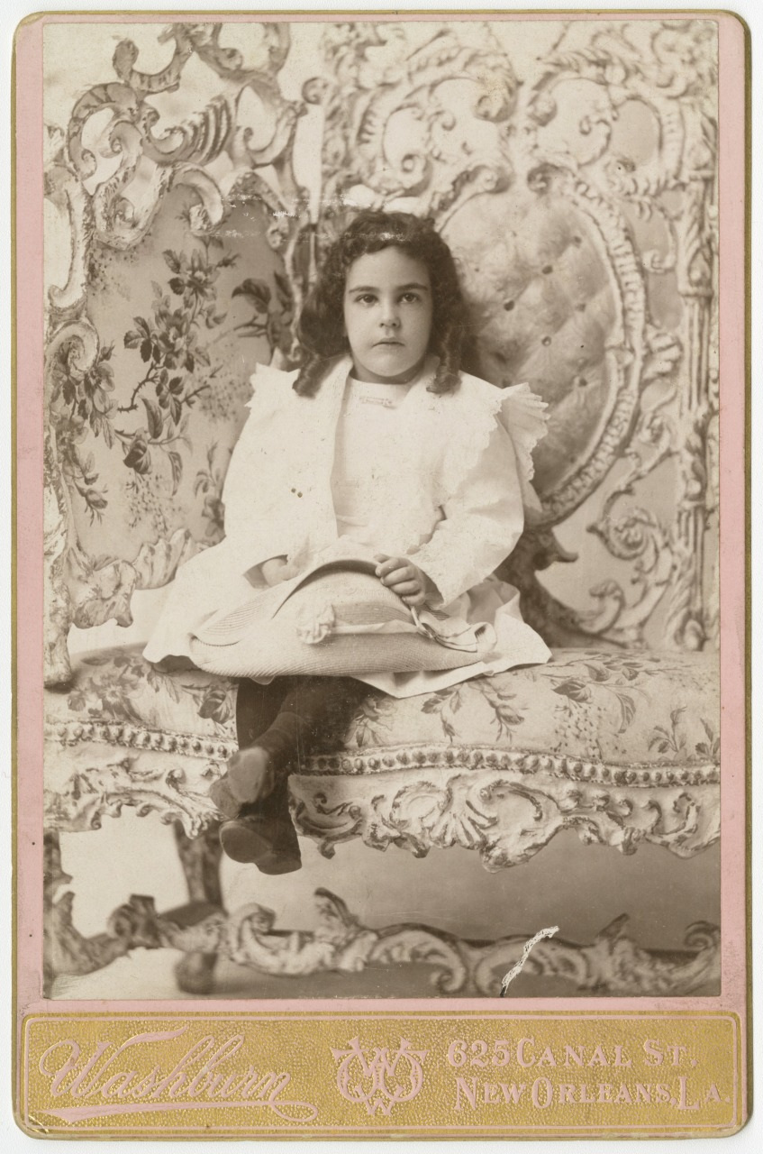 A vintage photograph of a young girl sitting on an ornate chair. She wears a light-colored dress and dark stockings, with curly hair. The background features intricate, floral upholstery. The photo has a Washburn studio label from New Orleans.