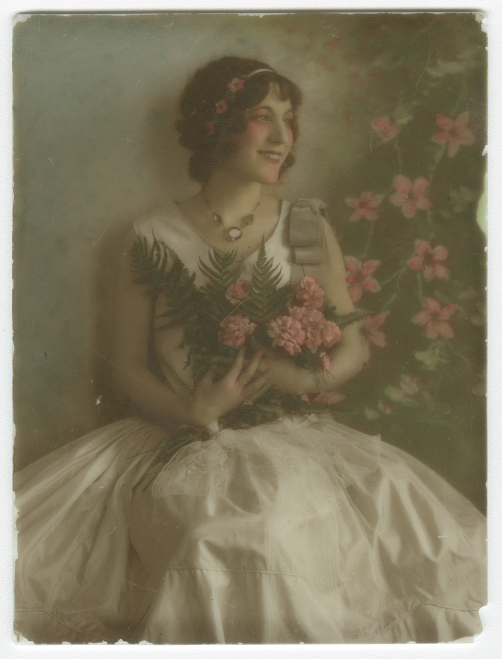 A woman in a vintage dress sits against a floral backdrop, holding pink flowers and ferns. She smiles softly, with small flowers adorning her hair. The image has a soft, nostalgic tone.