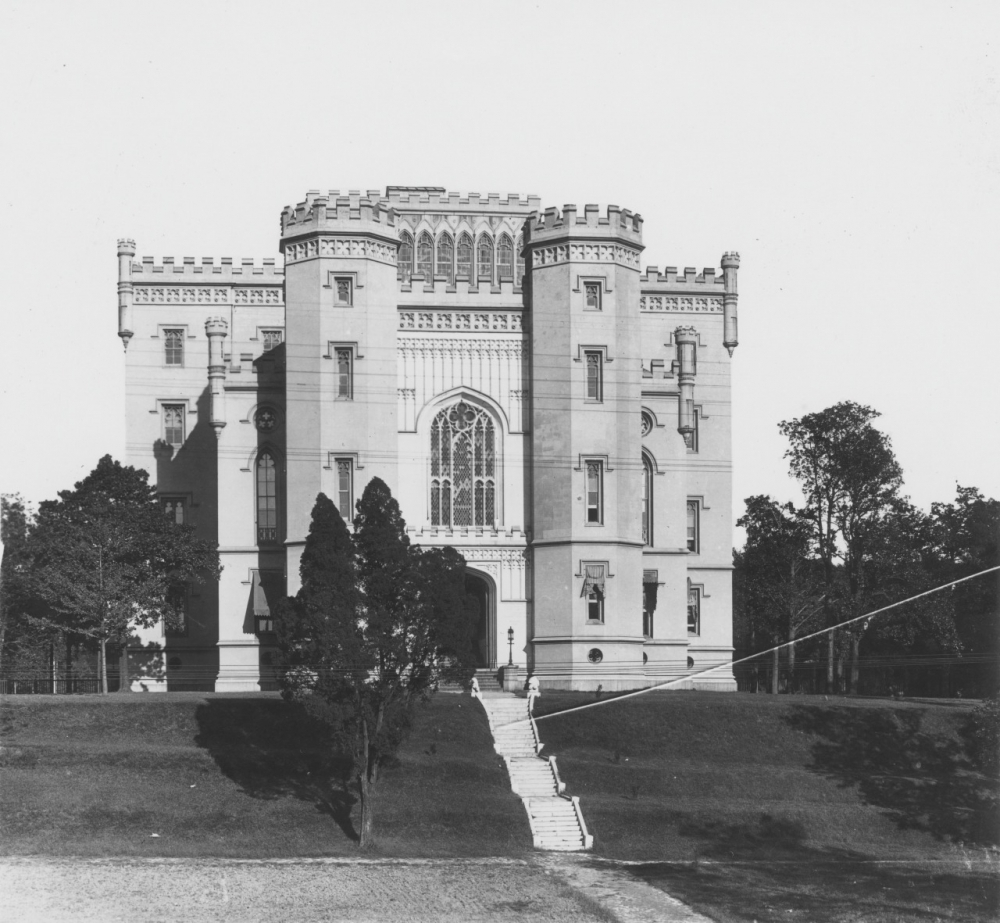 A historic Gothic-style building with a castle-like appearance stands amid trees. The facade features tall towers and arched windows, with a staircase leading up to the entrance. The photo is in black and white, highlighting architectural details.