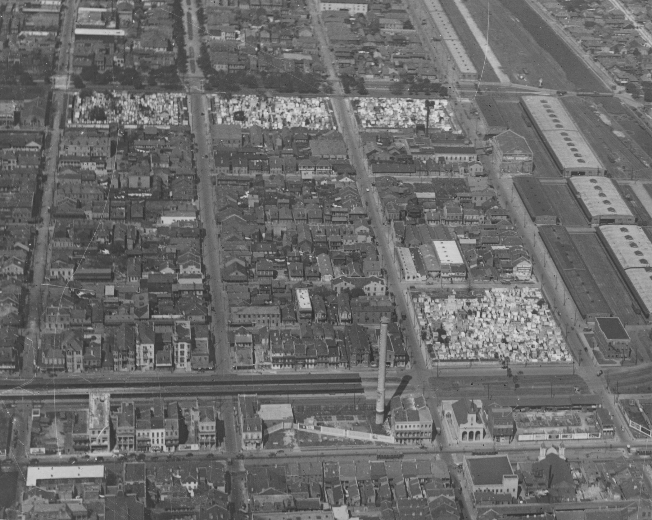 Aerial view of a densely built urban area with rows of houses and some larger industrial buildings. Streets are laid out in a grid pattern, and theres a large open area with a sparse arrangement of trees visible in the top section.