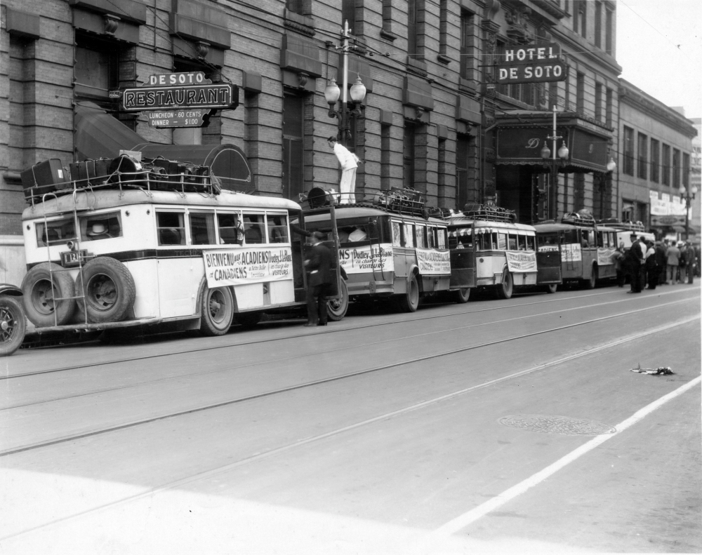 A row of vintage buses with luggage on top, parked in front of the Historic Hotel De Soto. The buses feature various travel advertisements and people are gathered nearby on the sidewalk.