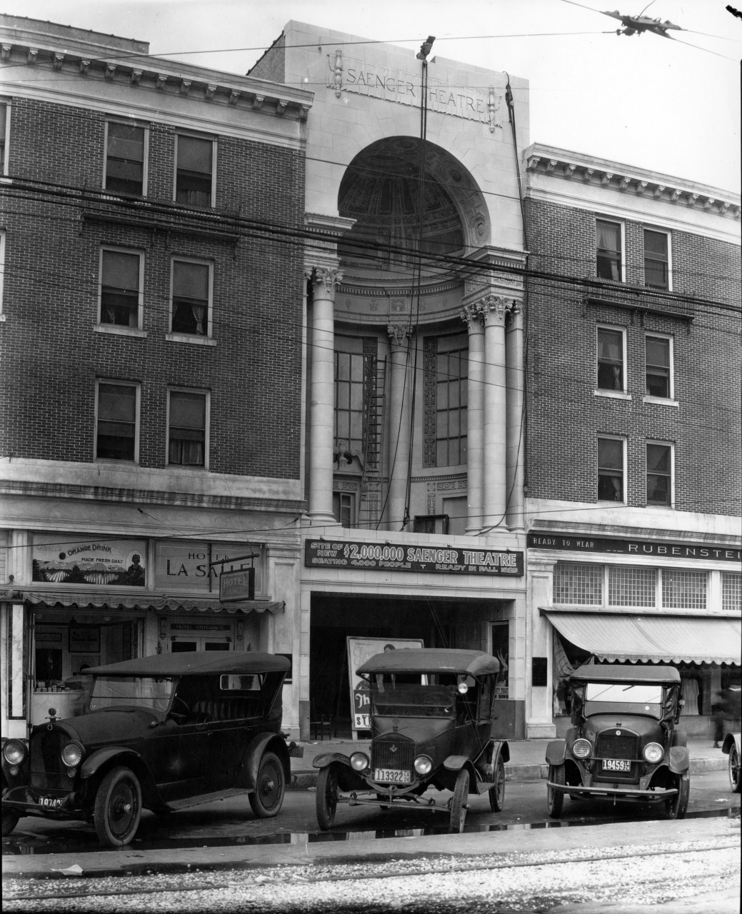Black and white photo of the Saenger Theatre with vintage cars parked in front. The building features a grand entrance with columns and ornate architectural details. The surrounding structures are brick with storefronts visible at street level.