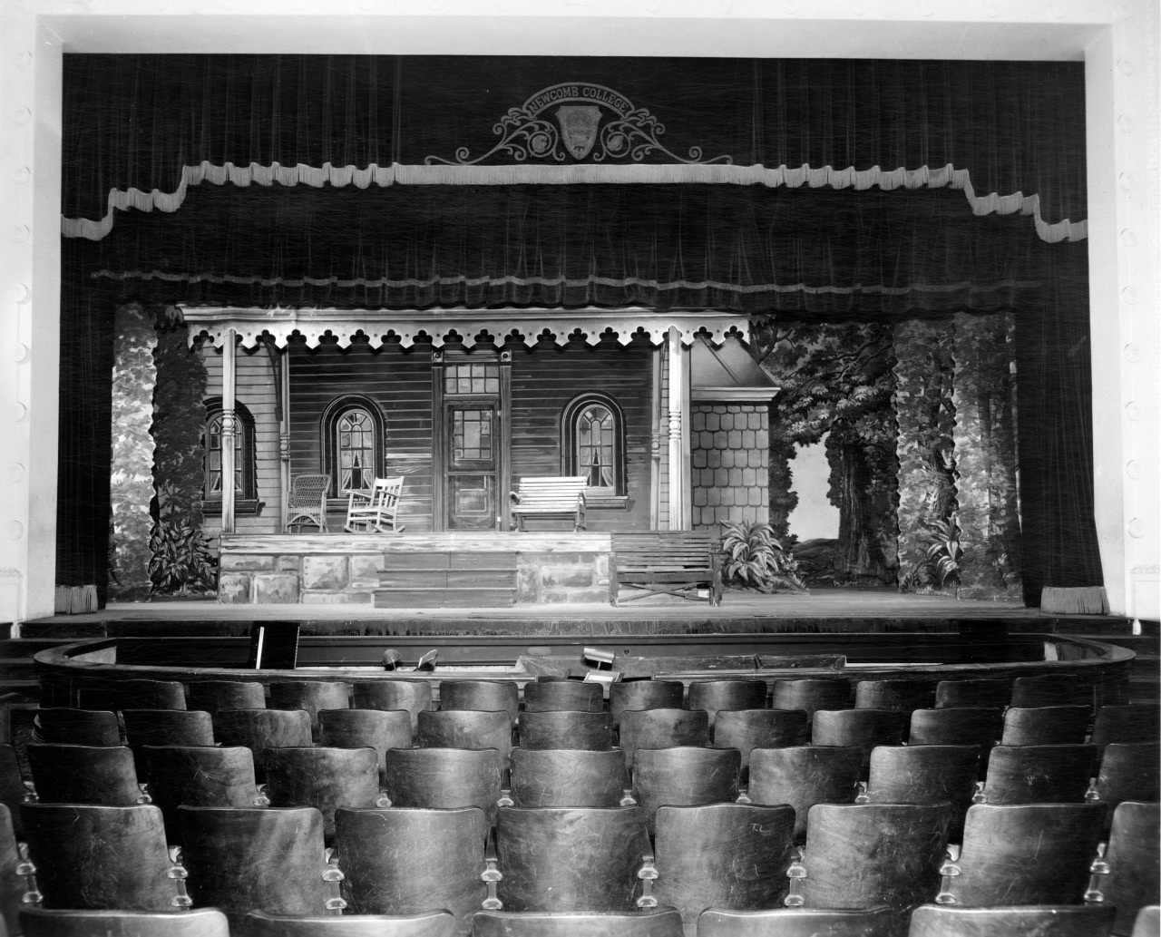 Black and white photo of a theater stage set. The scene includes a detailed wooden house façade with a porch, surrounded by trees and foliage. Empty theater seats are visible in the foreground.