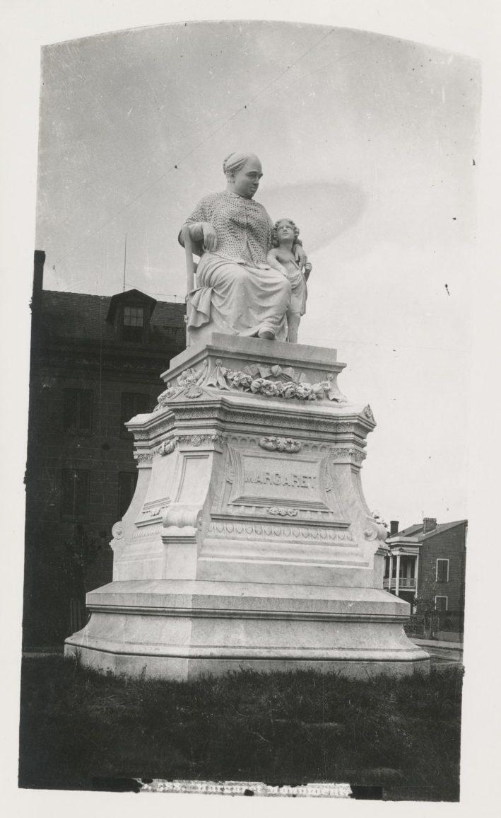 A black and white photo of a tall stone monument featuring a seated woman with a child at her side. The base of the monument has the name Margaret engraved on it. The monument is set on grass, with buildings visible in the background.