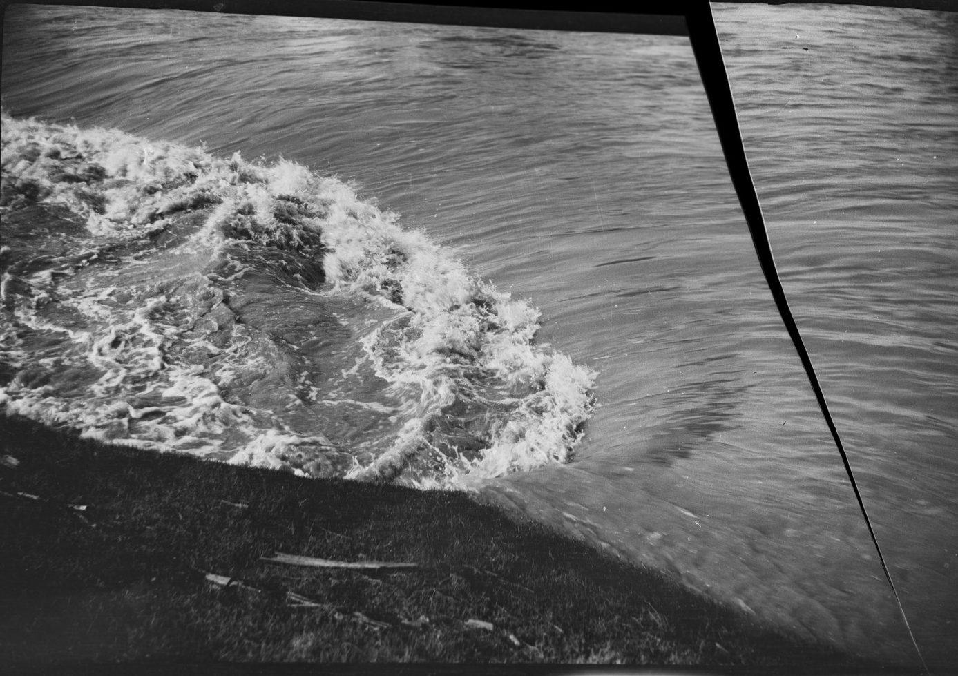 A black and white photograph of ocean waves crashing onto the shore, with a visible tear or fault line running vertically through the image. The shoreline is grassy, and the water appears rough with white foamy waves.