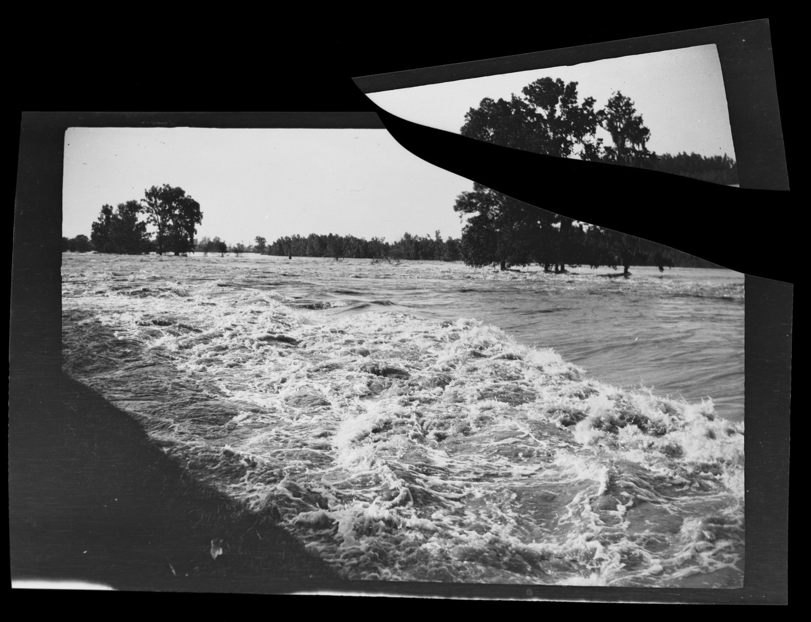 Black and white photo showing a flooded river with turbulent water flowing over a wide area. Trees stand partially submerged in the background. A portion of the image is torn and overlapped onto the main photo.