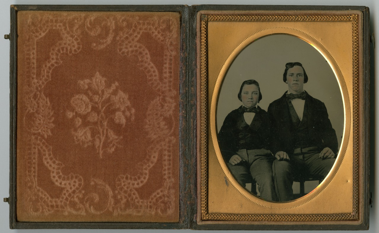 A vintage photograph in an ornate case showing two seated individuals in formal attire. The interior features a decorative red velvet lining on the left side and the photo on the right side, framed with intricate gold detailing.