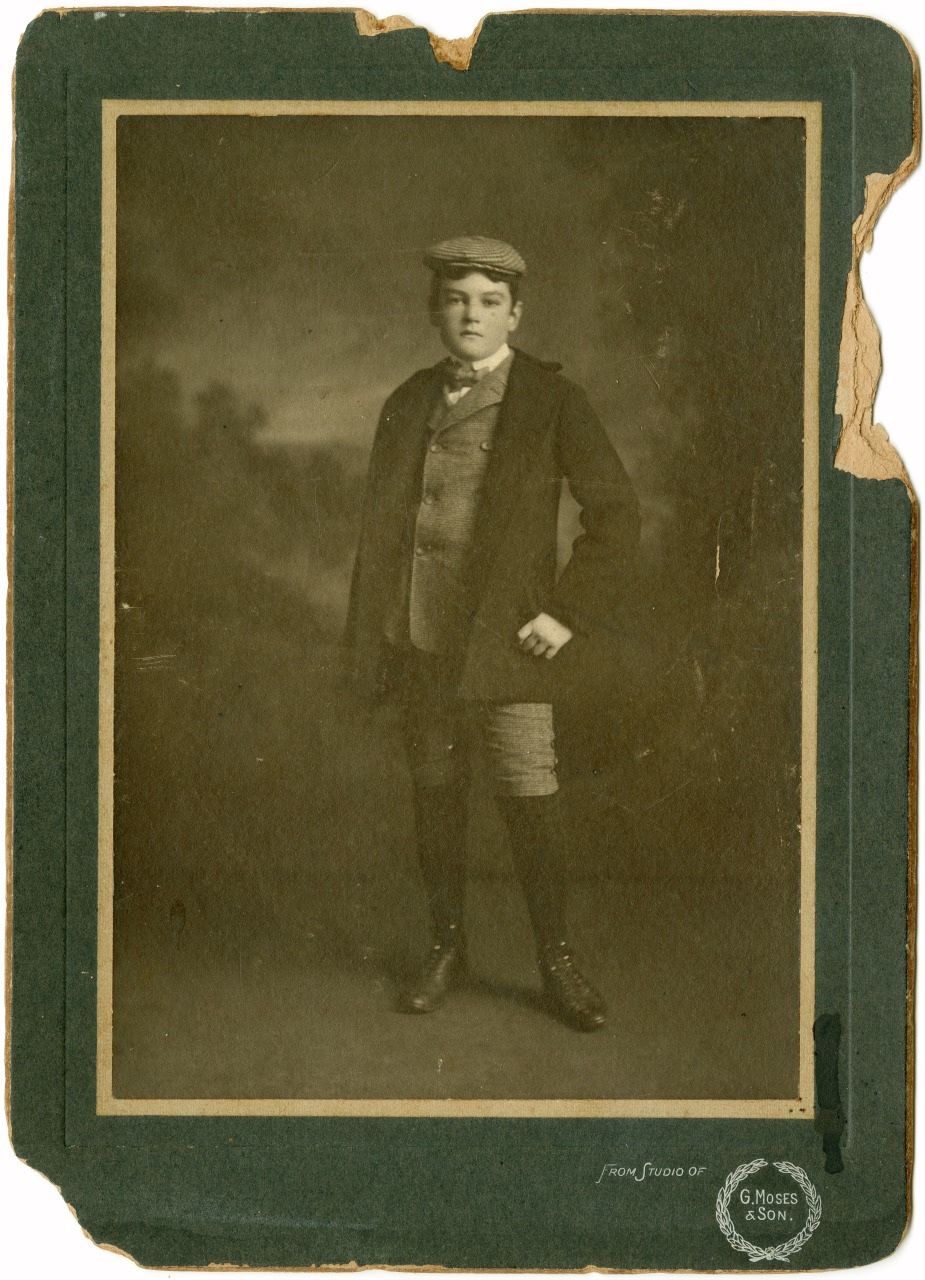 A vintage sepia-toned portrait of a young boy in a suit with knickerbockers, standing against a studio backdrop. The photograph shows signs of wear, with frayed edges and noticeable damage at the top right corner.