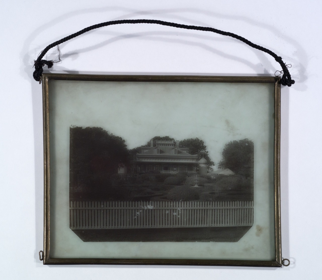A vintage framed photograph of a house, featuring a fence in the foreground and trees on both sides. The frame has a black hanging cord attached.