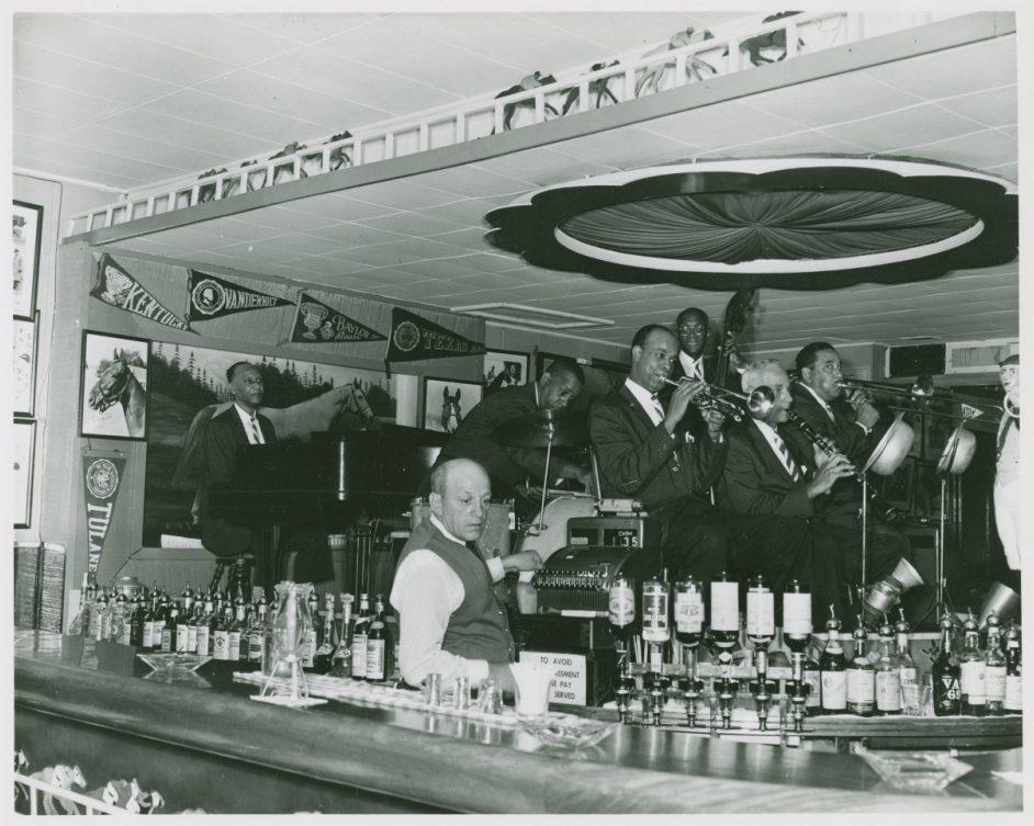 A jazz band performs in a lively bar. Musicians play trombone, trumpet, and saxophone beside a pianist. The bartender stands in front of a counter lined with bottles. Vintage posters decorate the walls under a ceiling fan.