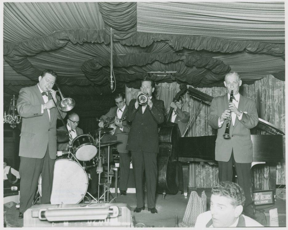 A black and white photo of a jazz band performing on stage. Musicians play trumpet, clarinet, and trombone, with a pianist, drummer, and double bassist in the background. The stage is decorated with draped fabric. Audience members are partially visible.
