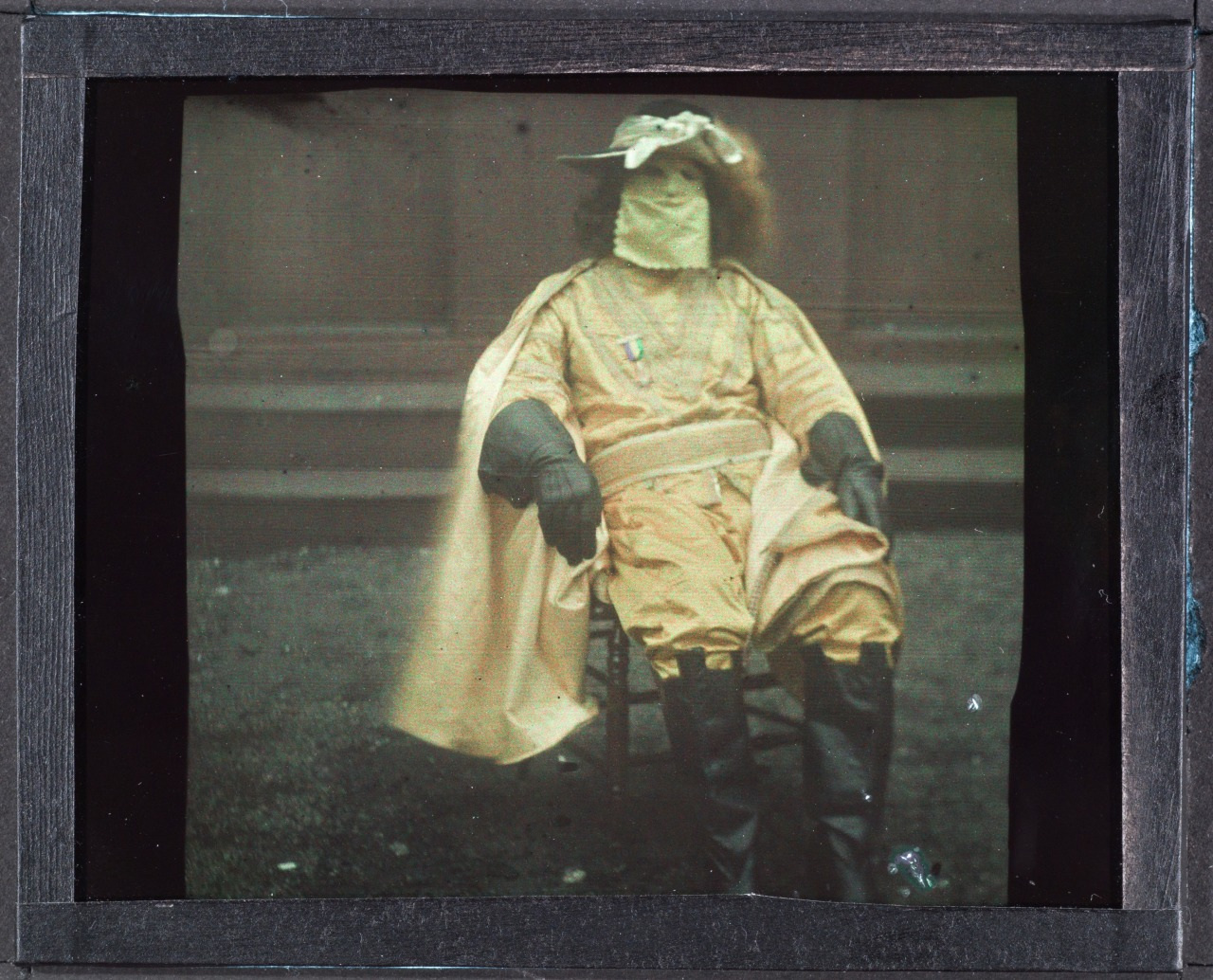 A person wearing a yellow costume with a covered face and large gloves sits on a chair outside. The outfit includes a decorative hat with feathers. The background appears to be a wooden structure.
