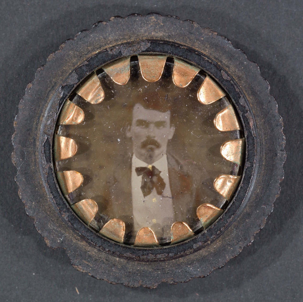 A vintage circular photograph featuring a man with a mustache, wearing a suit with a bow tie. The photo is framed within a gear-like metal border. The image is worn and faded, giving it an antique appearance.