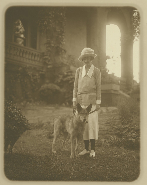 A sepia-toned photograph of a woman in a long dress and hat standing beside a wolf-like dog. They are in front of a grand building with a large arched window and surrounded by lush greenery.