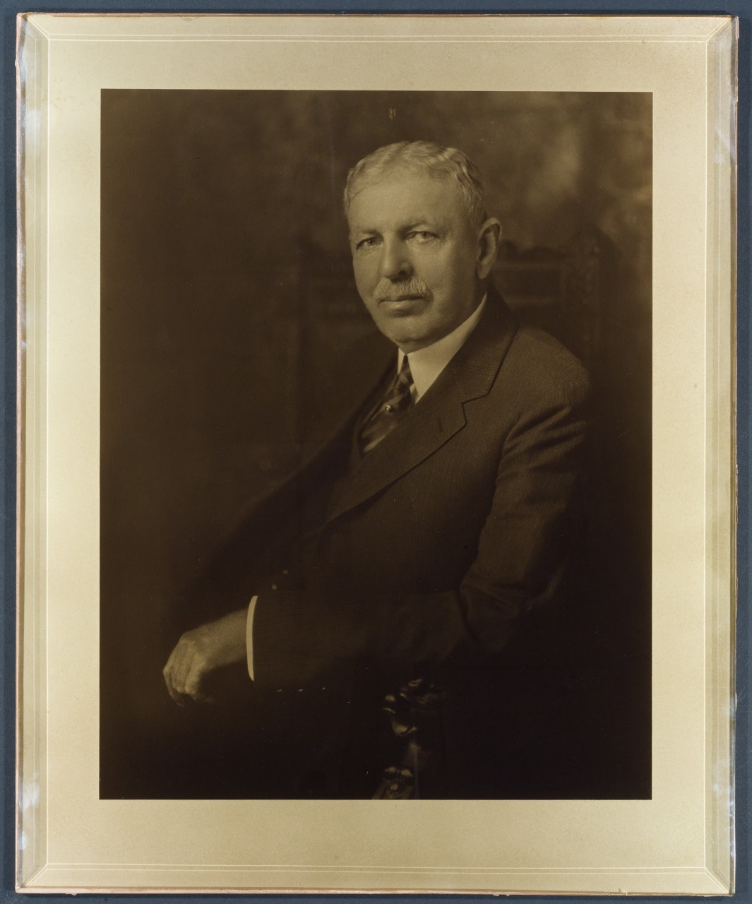 Sepia-toned portrait of an older man wearing a suit, seated with one arm on a chair. He has short hair and a mustache, and hes looking slightly to his left. The background is blurred, and the photo has a vintage, framed appearance.
