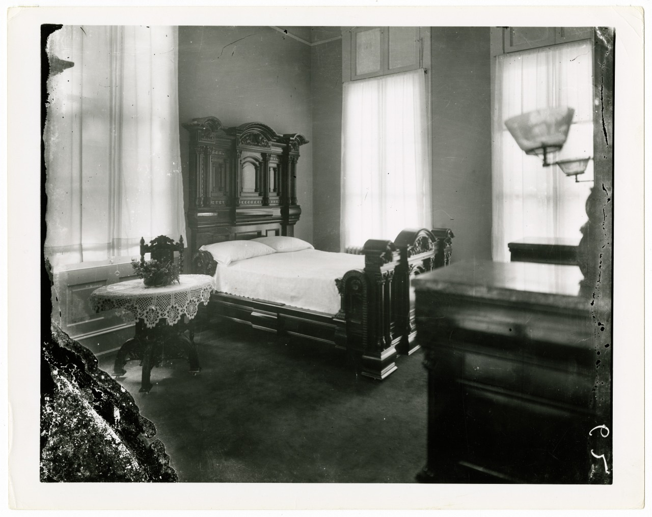 A vintage black and white photo of a bedroom with an ornate wooden bed, lace-covered round table, and antique furniture. Large windows with sheer curtains illuminate the room, creating a nostalgic and elegant atmosphere.