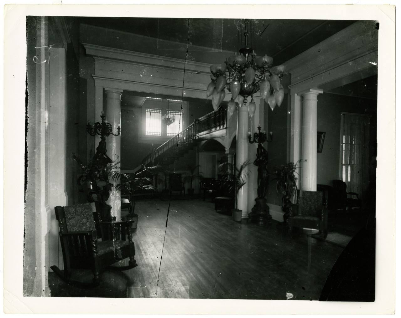 A vintage black and white photo of an elegant interior with a grand staircase, ornate columns, and an elaborate chandelier. Rocking chairs and potted plants are arranged along the wooden floor, creating a classic, inviting ambiance.