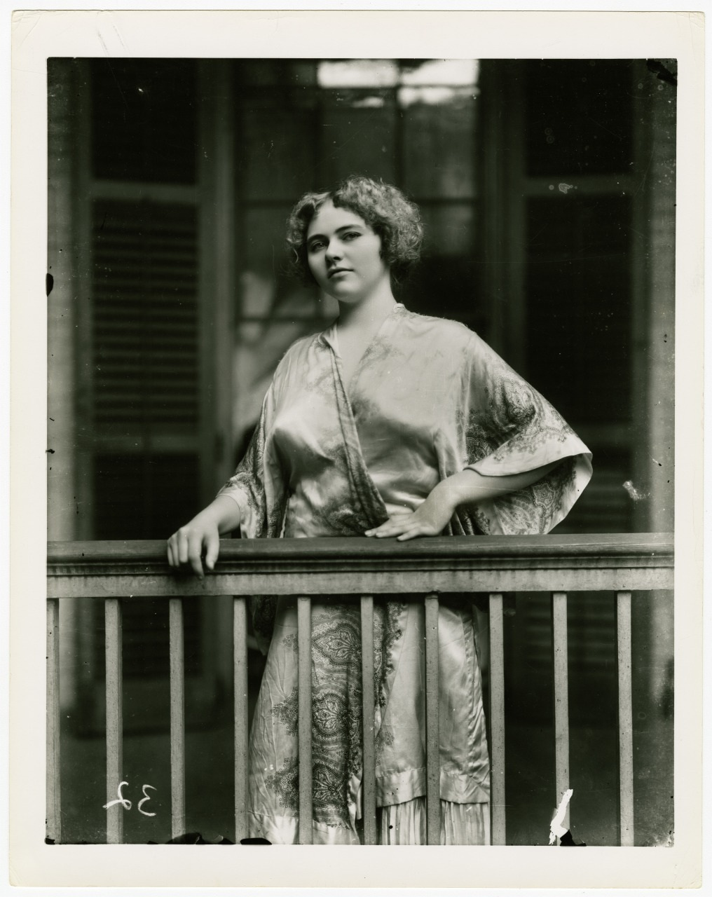 A woman in a vintage silk robe with intricate patterns poses gracefully, leaning on a wooden railing. Her expression is confident and serene, surrounded by blurred architectural details in the background.