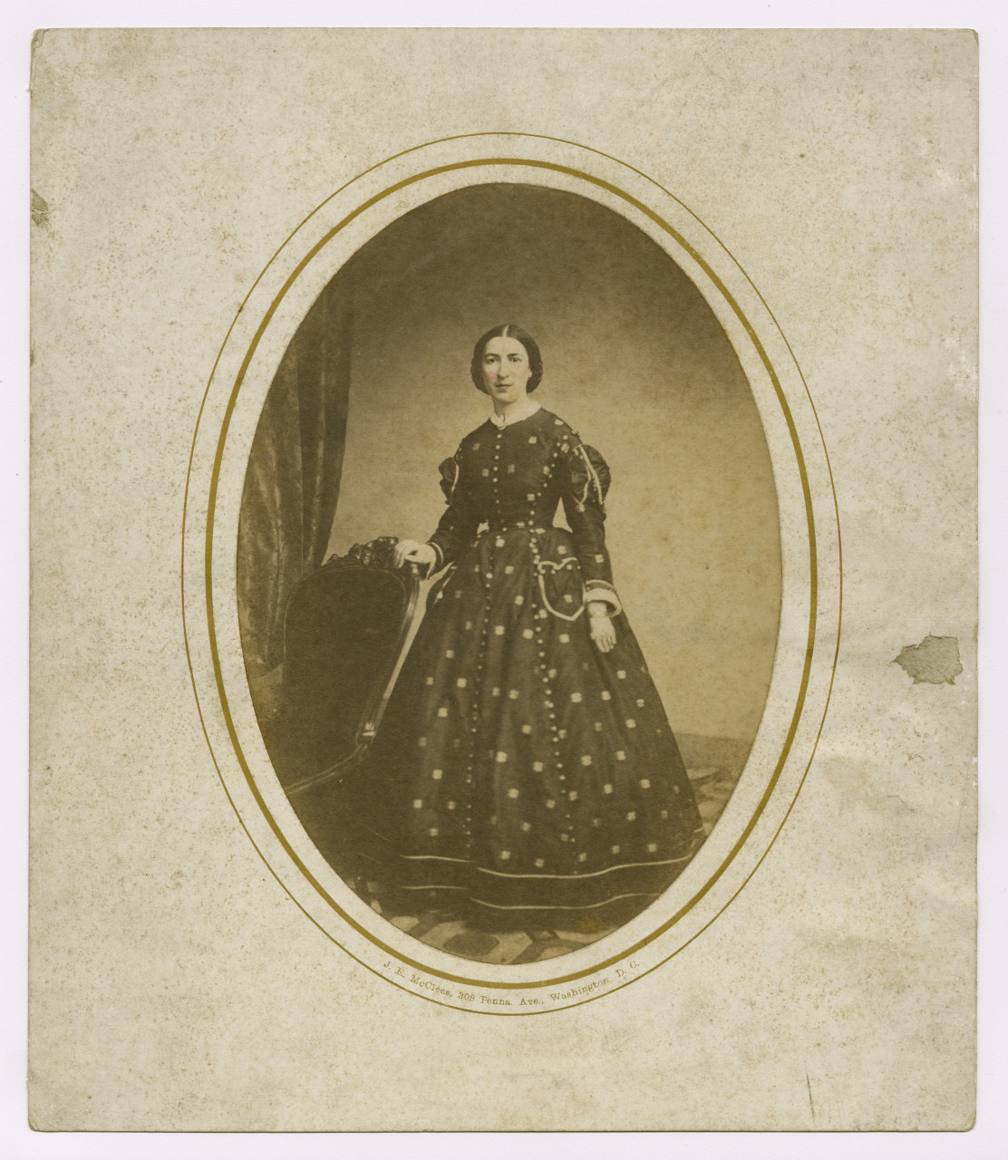 A sepia-toned vintage portrait of a woman in a 19th-century dress. She stands with one hand resting on a chair, framed by an oval border. The photo has aged marks and is bordered by a plain type of mat.