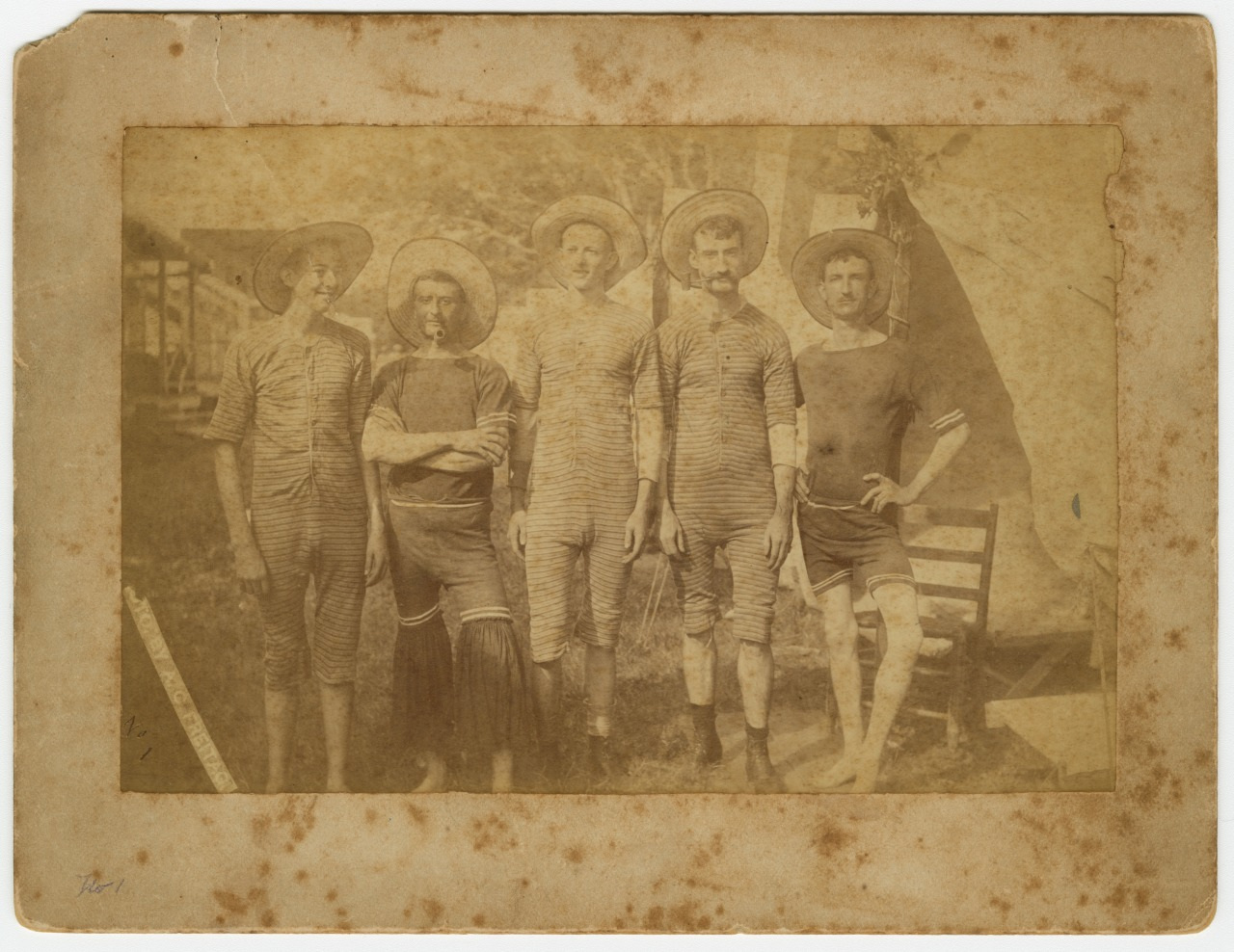 A vintage sepia-toned photograph of five men wearing striped swimsuits and straw hats. They pose casually outdoors, with trees and a wooden structure in the background. The image is aged, with visible marks and discoloration.