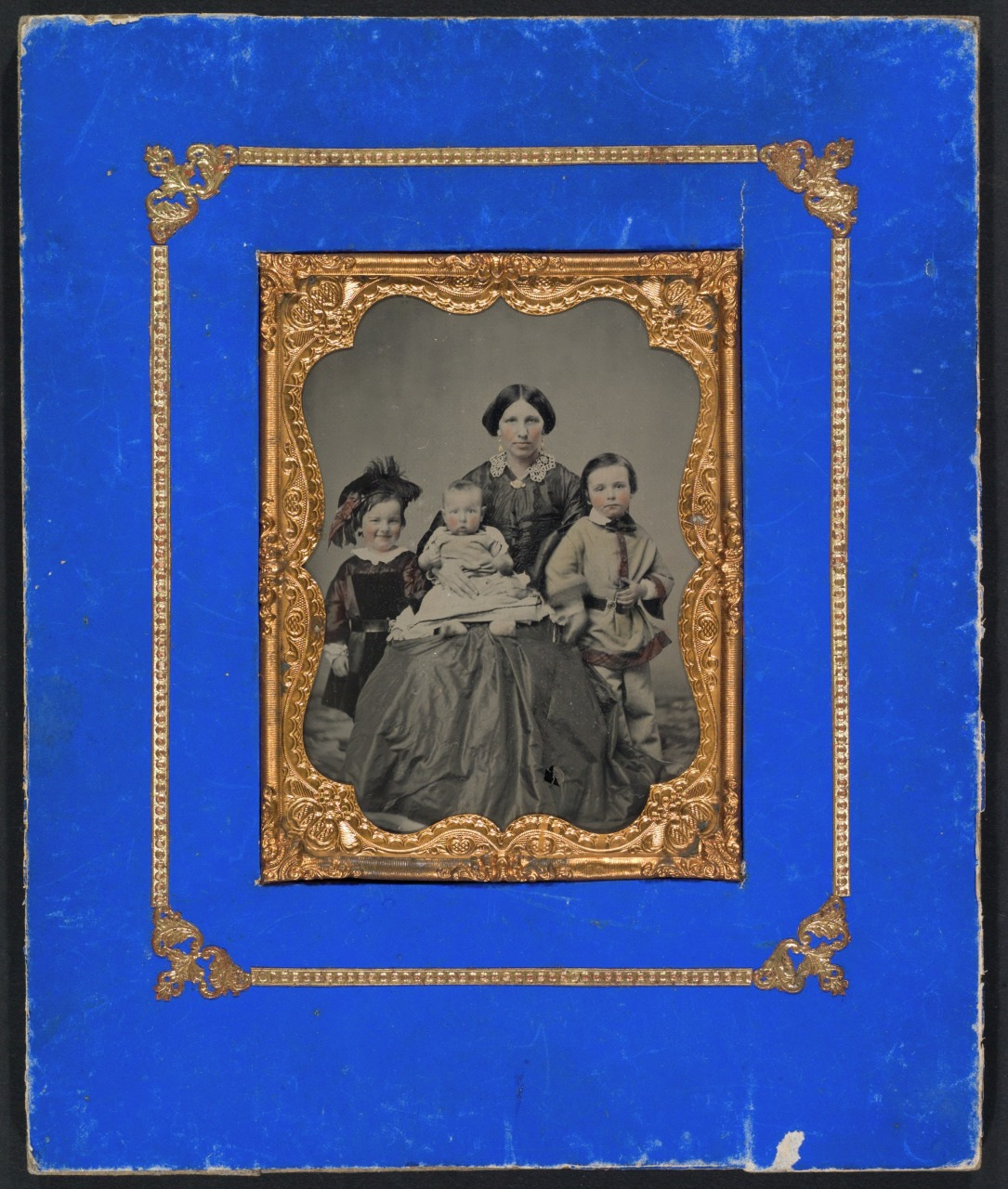 An old photograph with an ornate gold frame on a blue mat features a seated woman holding a baby, flanked by a young girl and a young boy standing beside her. They are dressed in period clothing.