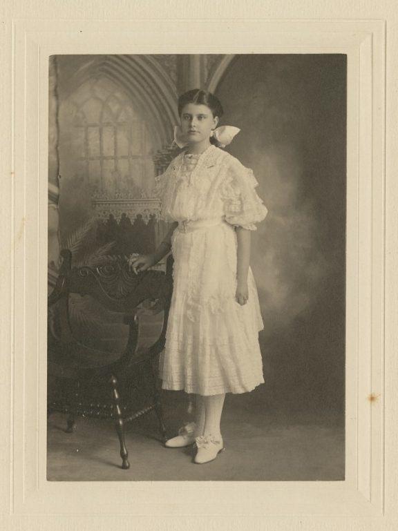 A vintage black and white photo of a young woman in a white Victorian dress standing beside an ornate wooden chair. She has a bow in her hair and is gazing directly at the camera. The backdrop features a gothic-style window design.