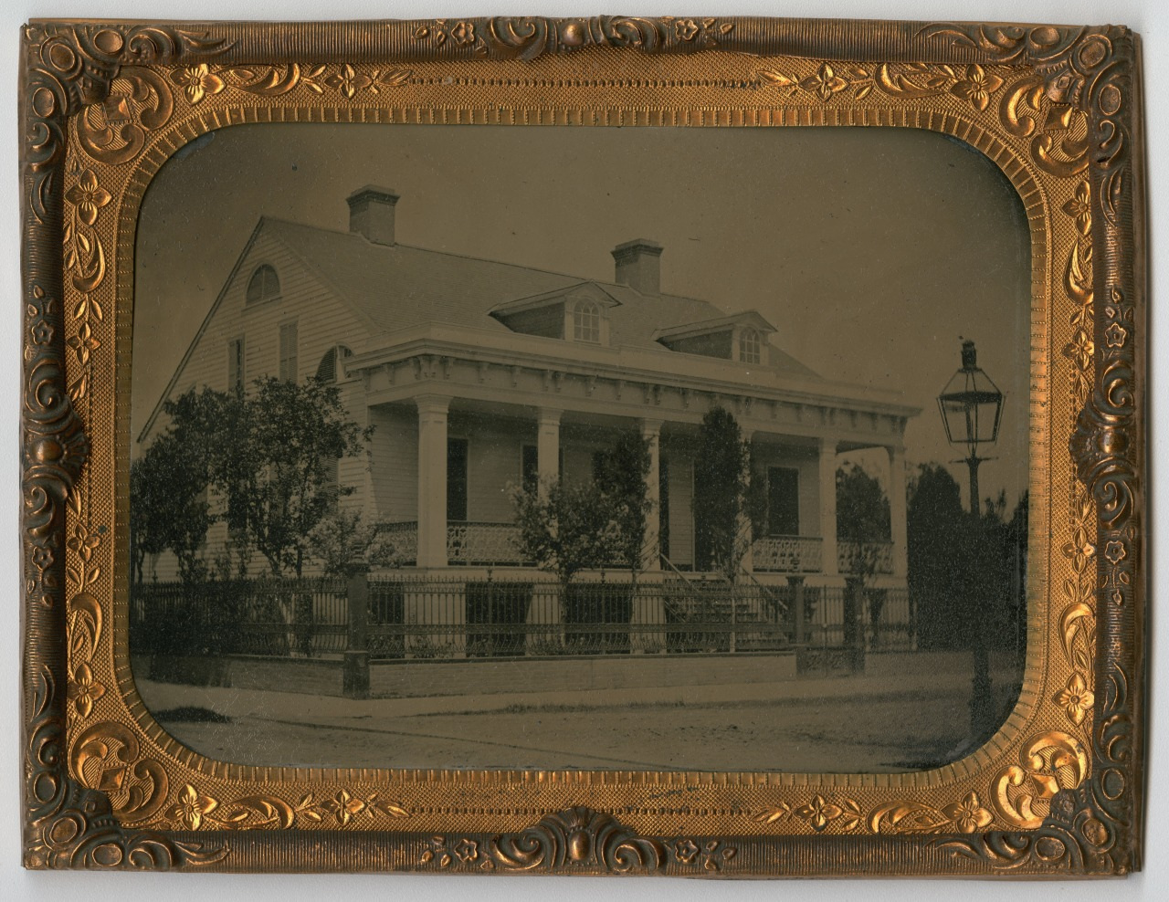 A vintage photograph of a large, two-story house with a covered porch, white columns, and dormer windows. It is surrounded by trees and enclosed by a decorative fence, all set in an ornate golden frame.