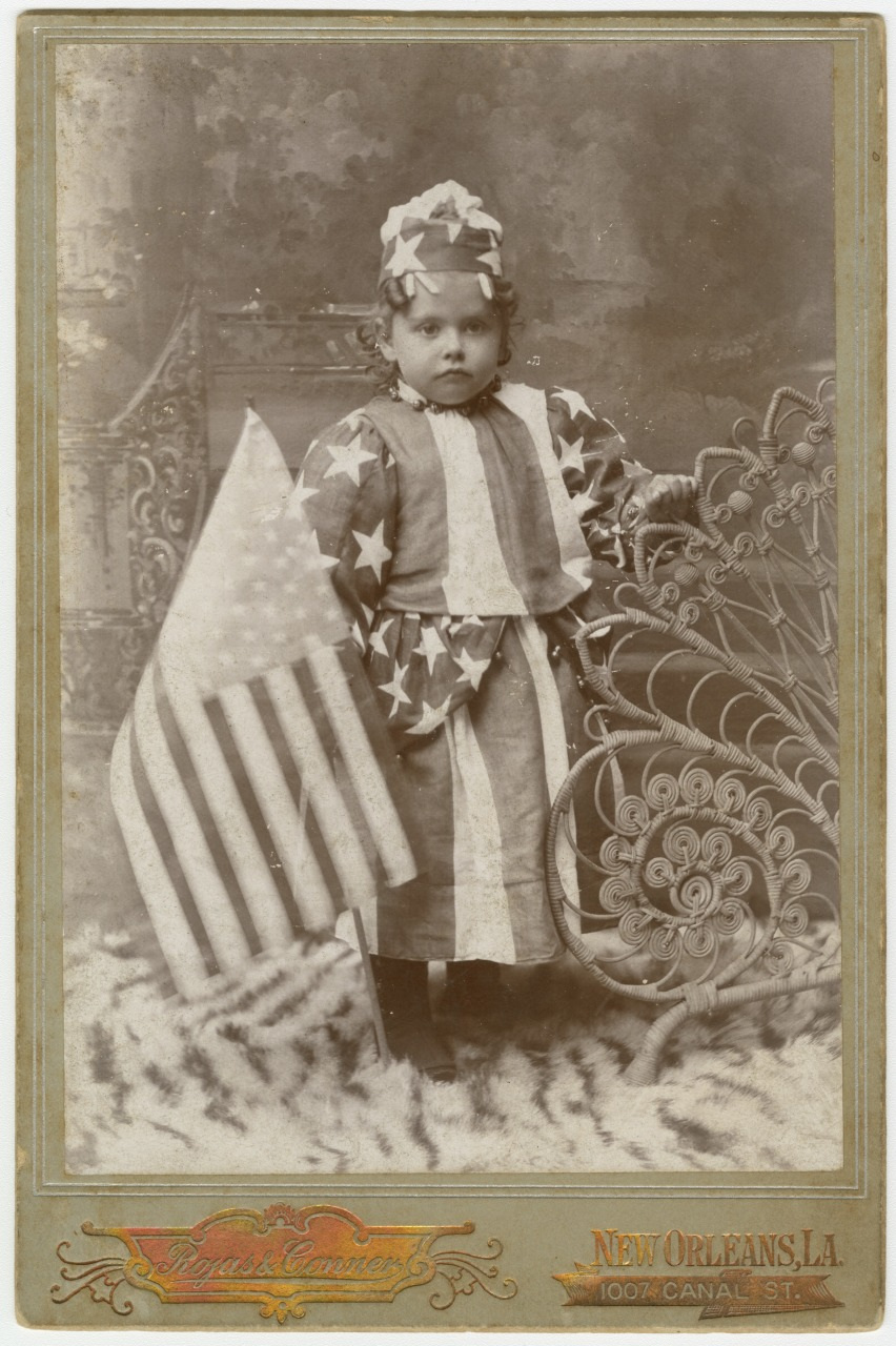 A sepia-toned vintage photograph of a young child dressed in a patriotic American flag-themed outfit, holding a large U.S. flag. The background features ornate decor, and the photo was taken in New Orleans, LA.