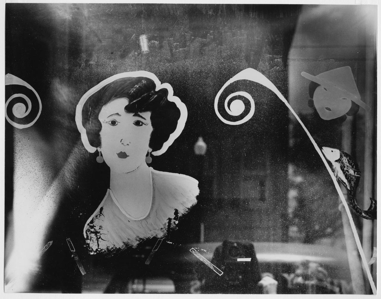 A window display features a painted portrait of a woman with dark hair and a serious expression. She is surrounded by decorative swirls and a stylized figure with a hat. The background is reflective, showing faint outlines of the street outside.