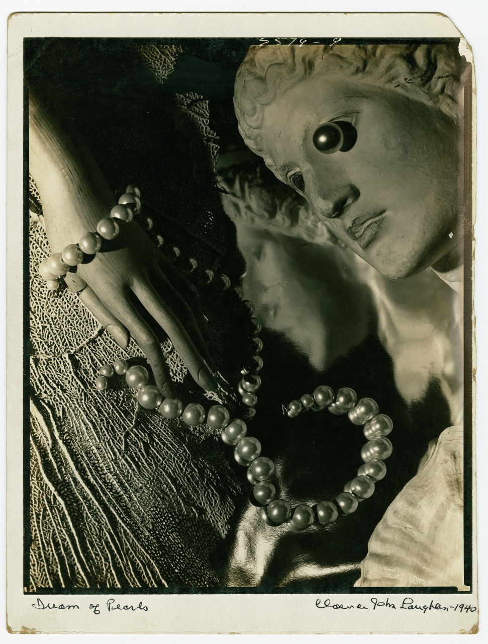 Black and white image titled Dream of Pearls by Clare Jota Laughar, 1940. A mannequin head and hand adorned with a pearl necklace rest on textured fabric, creating an ethereal, artistic composition.