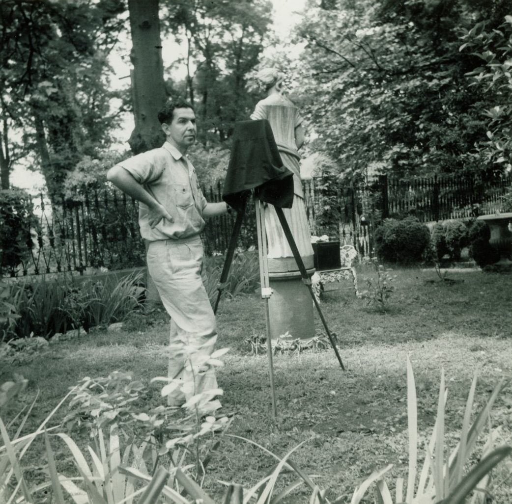 A man standing in a garden setting, adjusting a vintage camera on a tripod. Behind him is a statue surrounded by trees and plants. The scene suggests a moment captured during a photography session.