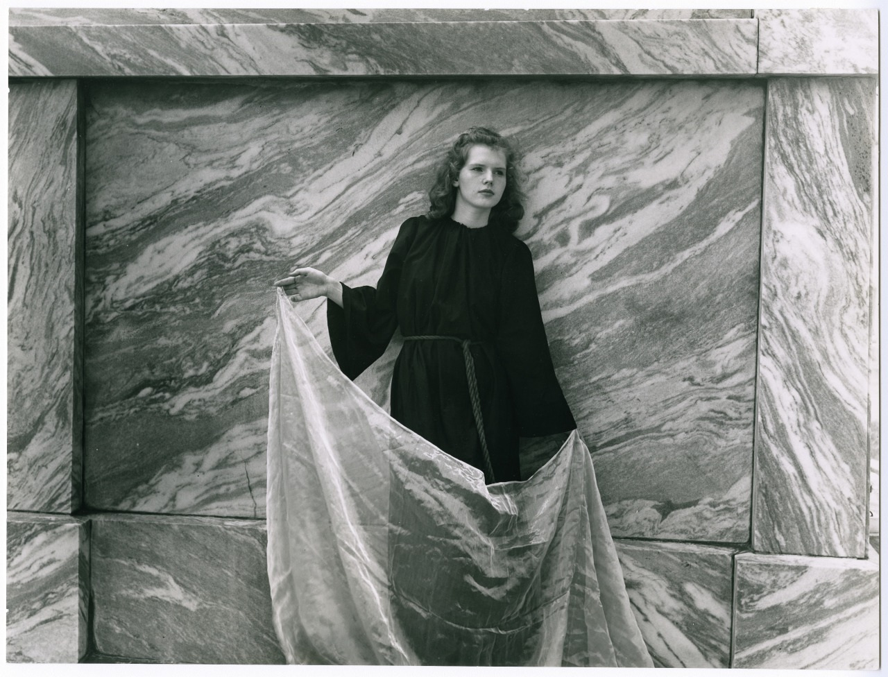 A woman in a dark dress poses against a marble wall, holding a sheer fabric. The textured wall has swirling patterns, and the fabric creates soft, flowing lines. The setting appears elegant and artistic.