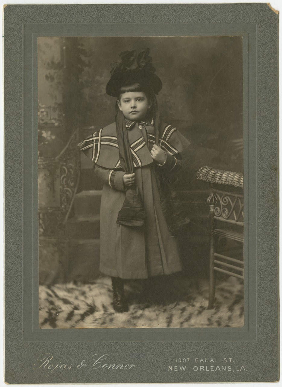 A vintage black-and-white portrait of a young child in a long coat and hat, standing next to a wicker chair. The backdrop is ornately decorated. The photograph is framed by a textured border with the studios name and address in New Orleans, LA.