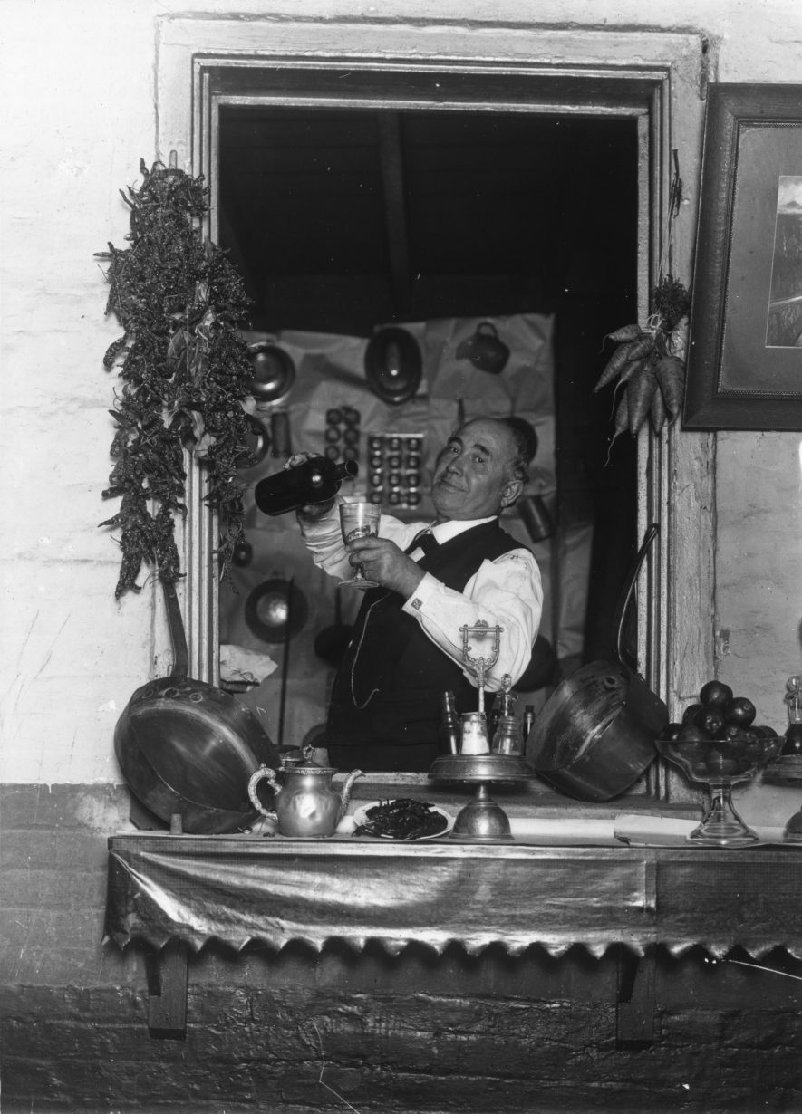 Monsieur Begue wears a vest and pours a bottle into a glass.