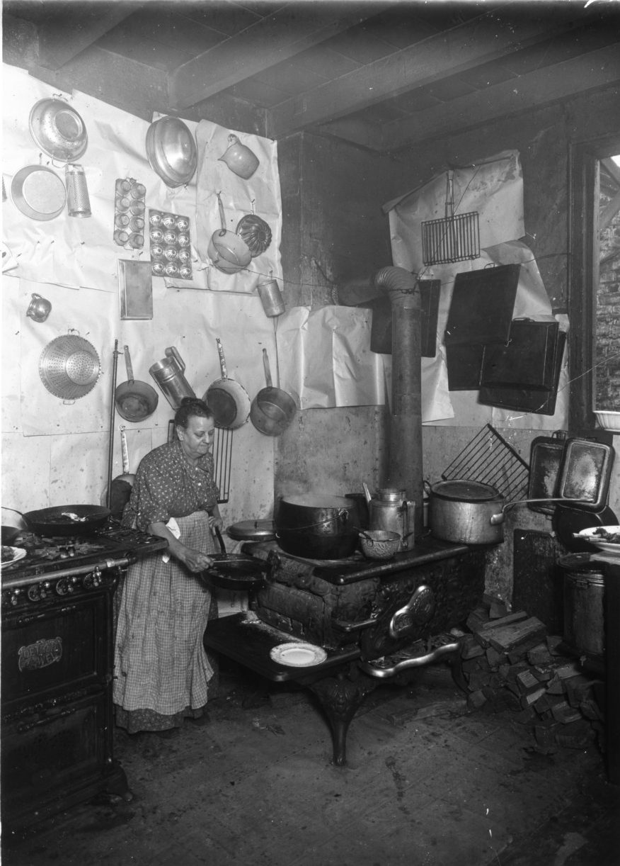 A black and white portrait of Madame Begue holding a frying pan in her kitchen near the stove.