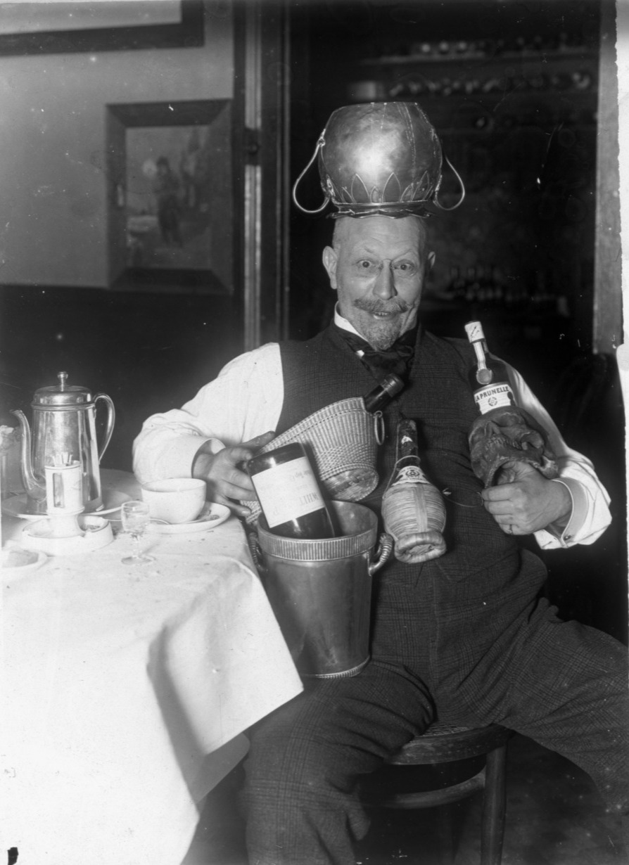 A man holds many bottles of wine and makes a crazed expression wearing an upside down carafe on his head.
