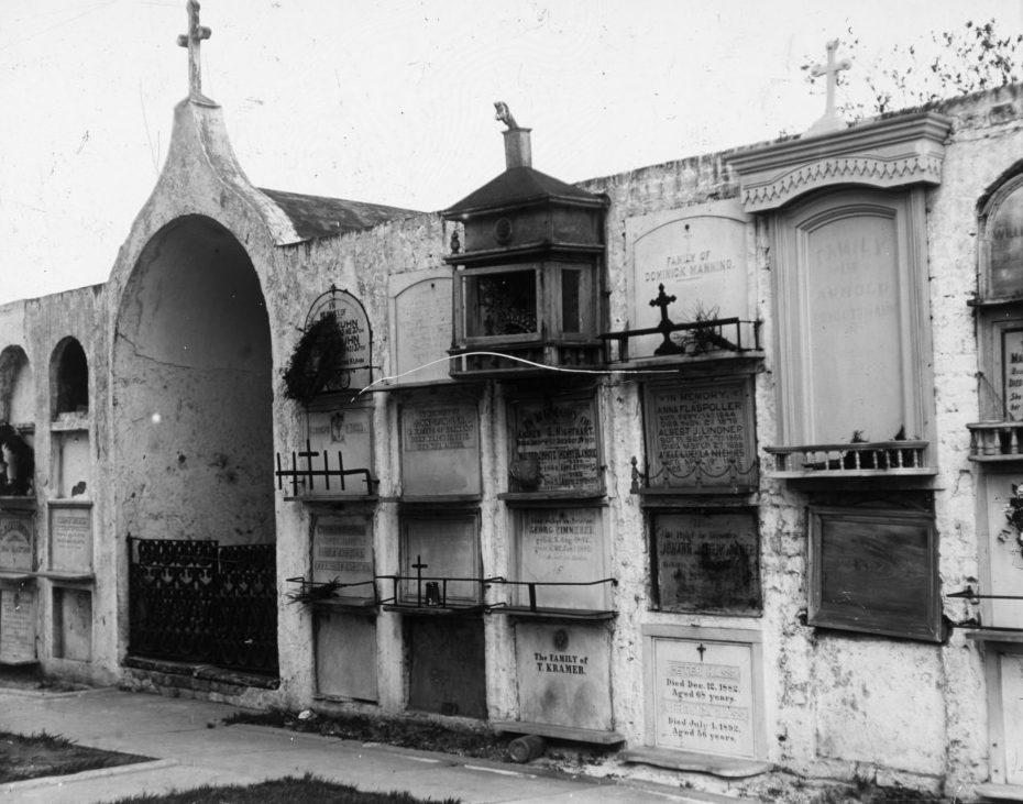 A black and white image of a cemetery wall with multiple tombs stacked vertically. Each tomb is marked with inscriptions, and some have small crosses. The wall shows signs of aging, with some greenery above.