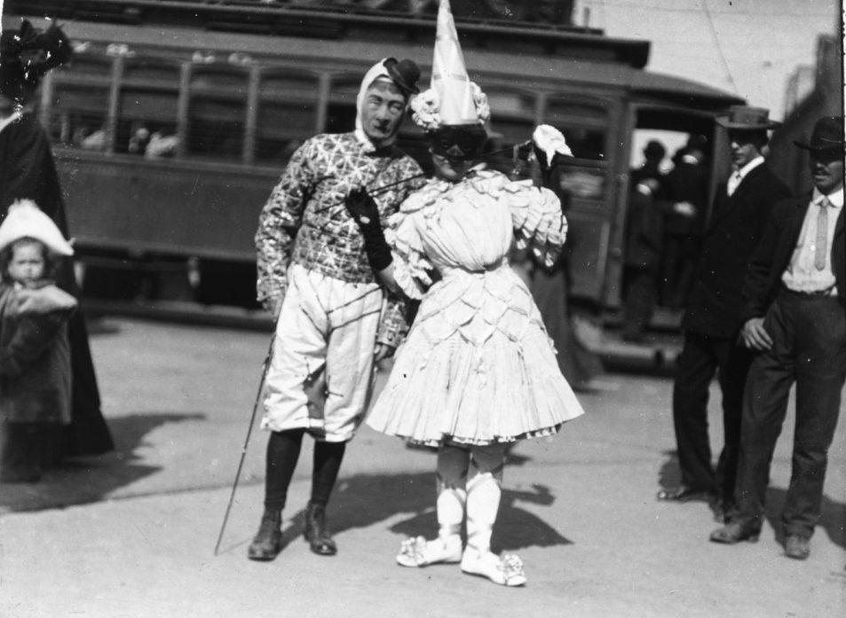 A couple in elaborate costumes from the early 1900s stand in front of a vintage streetcar. The man wears a patterned outfit and hat, holding a cane. The woman wears a frilly dress with a large hat. Bystanders and a child watch nearby.