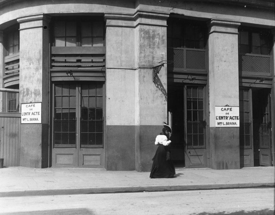 A woman in a long skirt and hat walks past a building with signs reading Café de lEntracte, Mme L. Brana. The façade features large windows and double doors with a small balcony above.