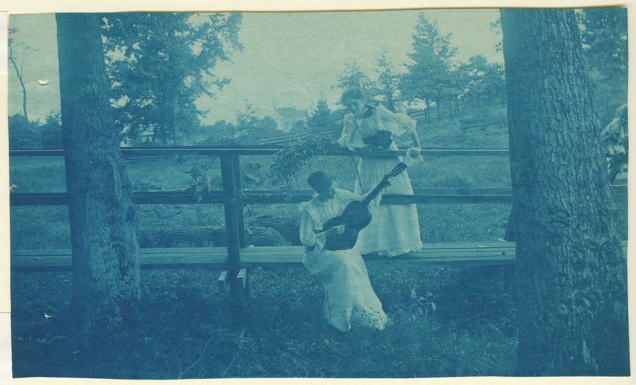 Two women in vintage dresses are outdoors by a wooden fence. One stands holding flowers, while the other sits playing a guitar. The background features trees under a cloudy sky. The image has a bluish tint, suggesting it is an old photograph.