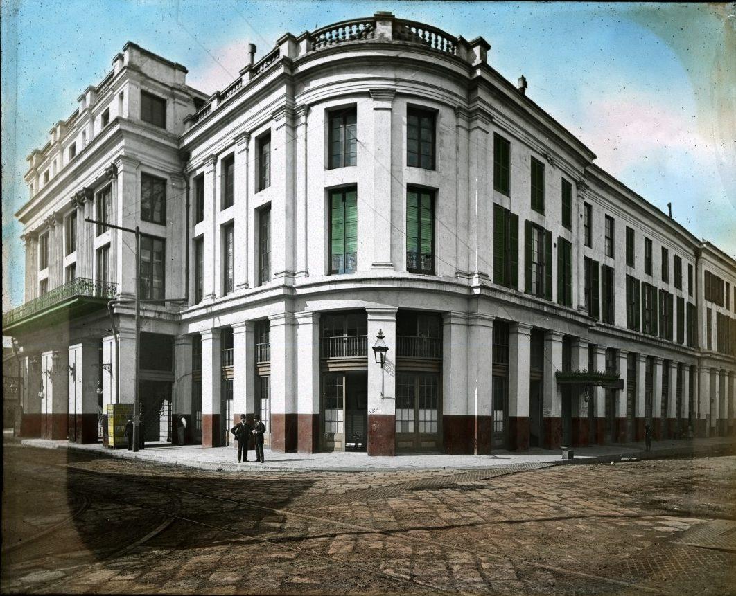A historic, multi-story building with large windows and classic architectural features is situated at a street corner. It has a curved facade and a few people are standing nearby on the cobblestone road under a clear sky.