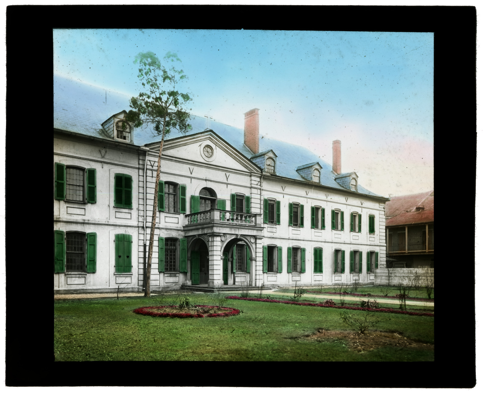 A historic white two-story building with green shutters and a central archway, set against a clear blue sky. The foreground features a well-kept lawn and flower beds arranged in neat patterns.