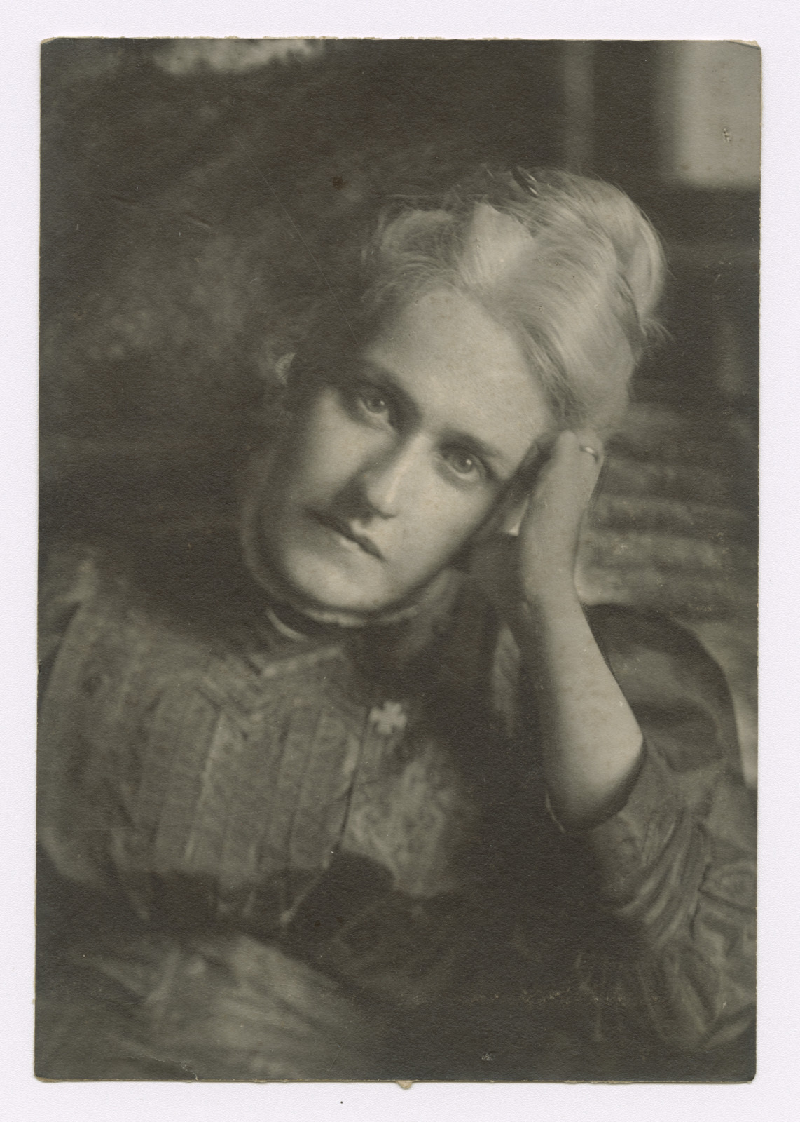 A vintage black and white photo of a woman with light-colored hair resting her head on her hand, wearing a high-collared, ornate dress. Her expression is thoughtful, and the background is softly blurred.